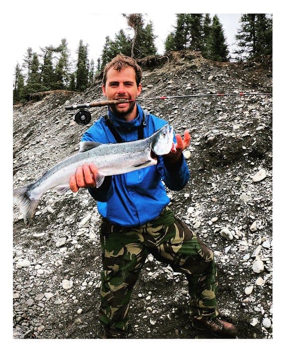 A polaroid: a man in a blue jacket holding up a freshly caught salmon on a riverbank
